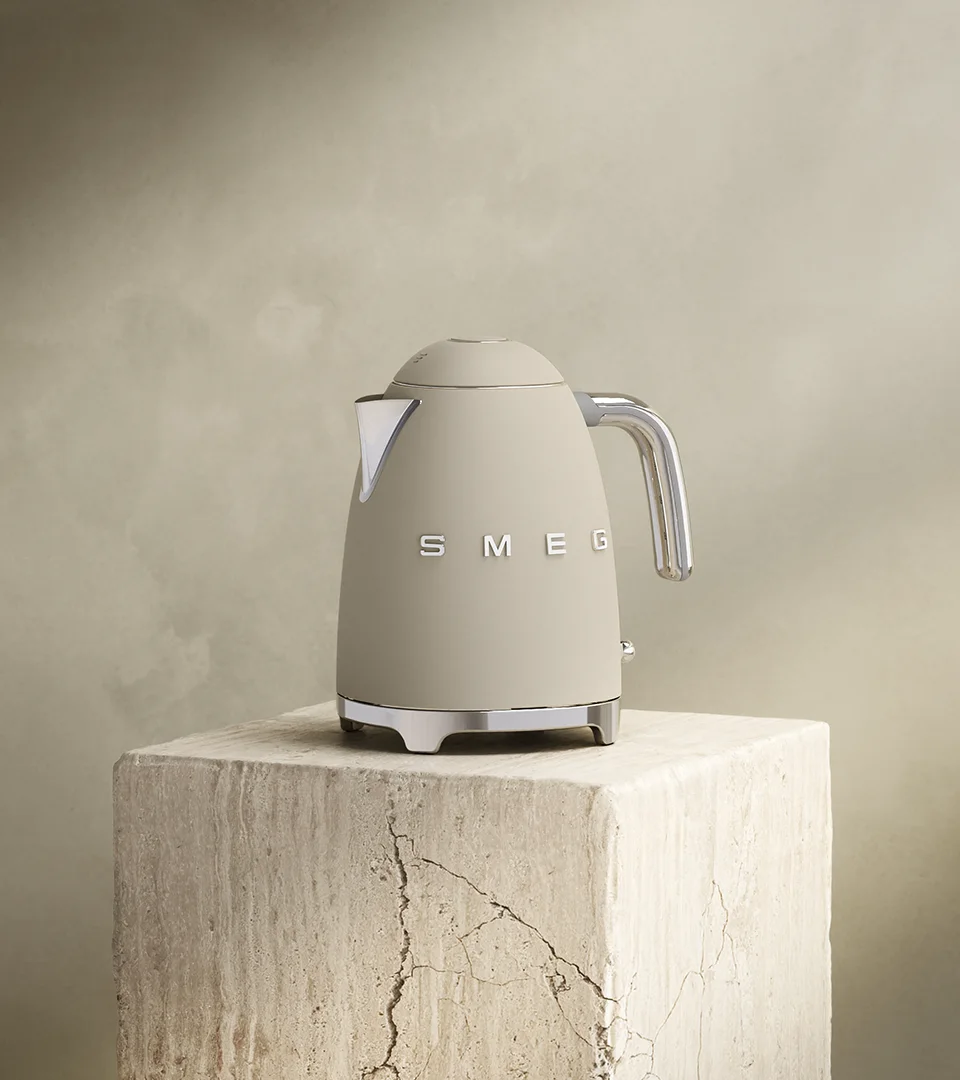 Matta beige Smeg-vedenkeitin, jossa kromikahva ja -jalusta, seisoo kuvioidulla beigellä lohkolla hillittyä taustaa vasten.