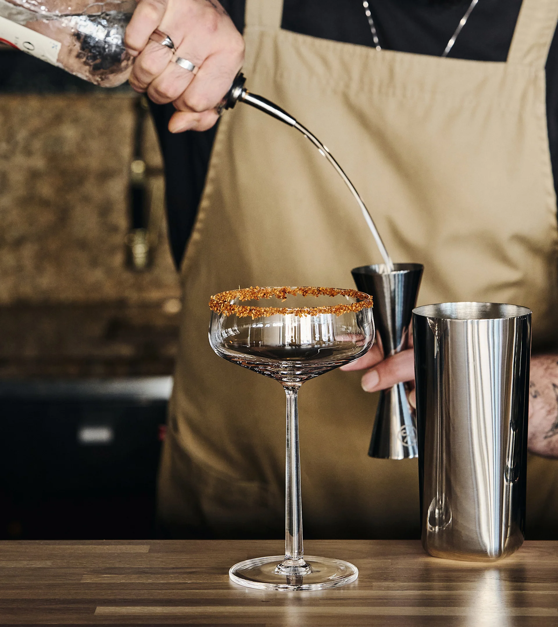Baarimikko kaataa juomaa pullosta mittalasiin, esillä sokerireunustettu cocktaillasi ja ravistin puupöydällä.