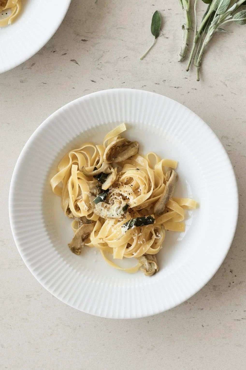 Pastaa artisokkien, salvia ja raastetun juuston kera valkoisella lautasella, salvianoksia vaalealla pöydällä.