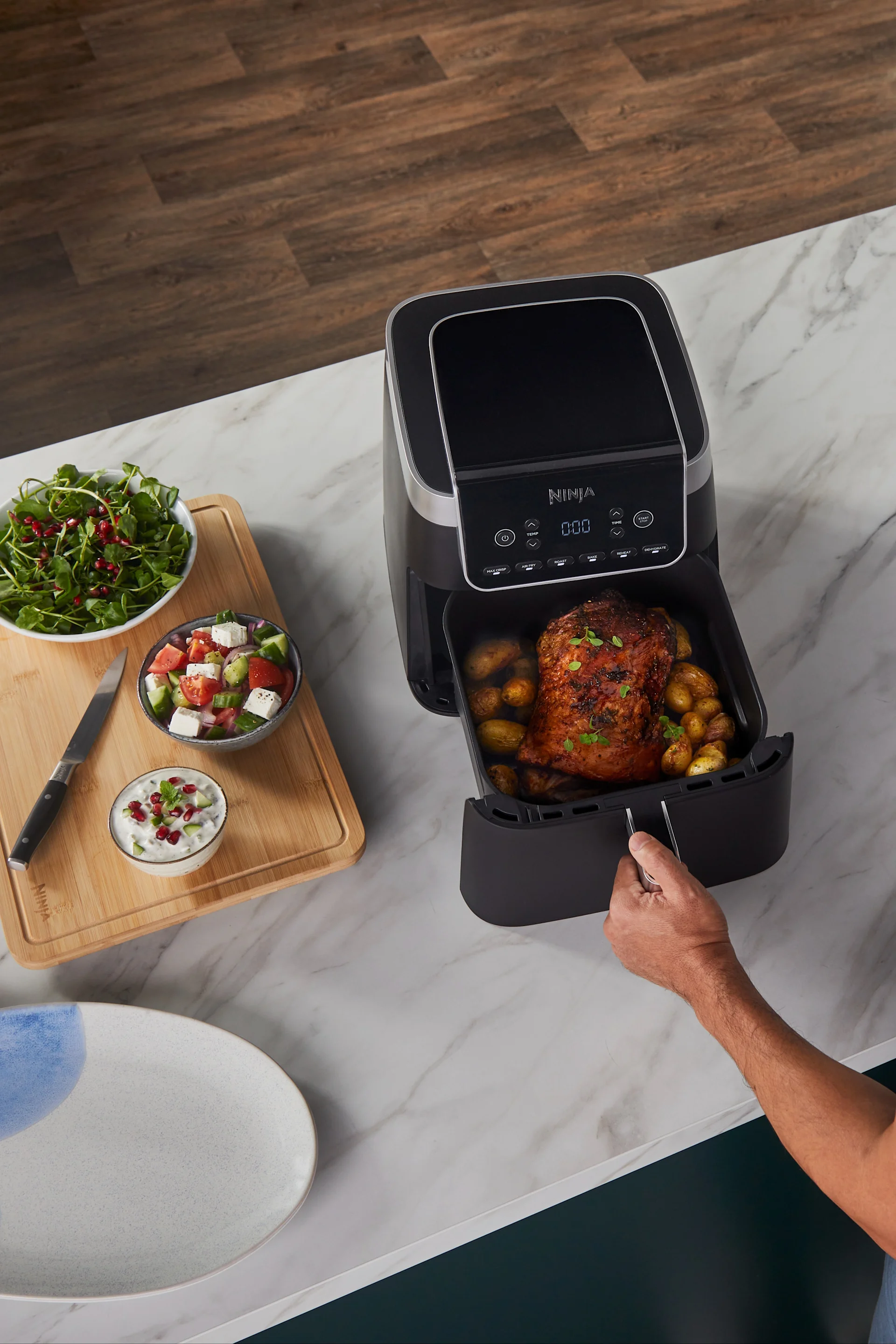 Ylhäältä kuva henkilöstä vetämässä mustaa airfryer-laatikkoa, jossa paahdettua kanaa ja perunoita, vieressä salaatteja ja dippiä.