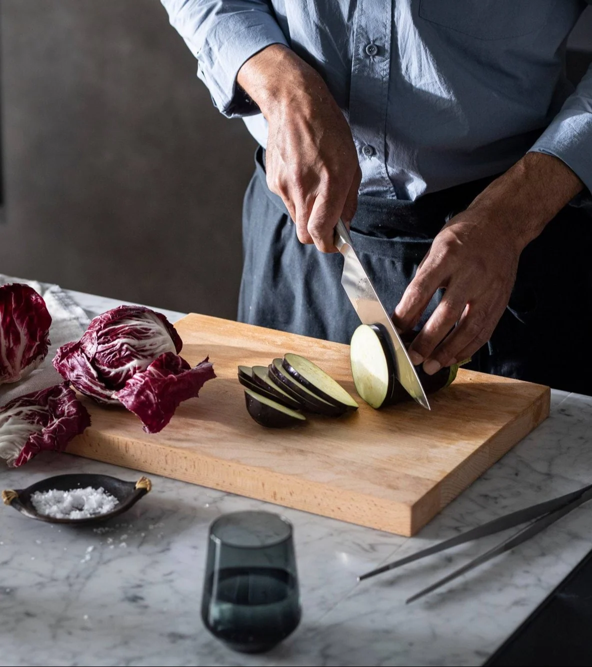 Lähikuva henkilöstä, joka viipaloi munakoisoa puisella leikkuulaudalla radicchio ja suola lähellä.