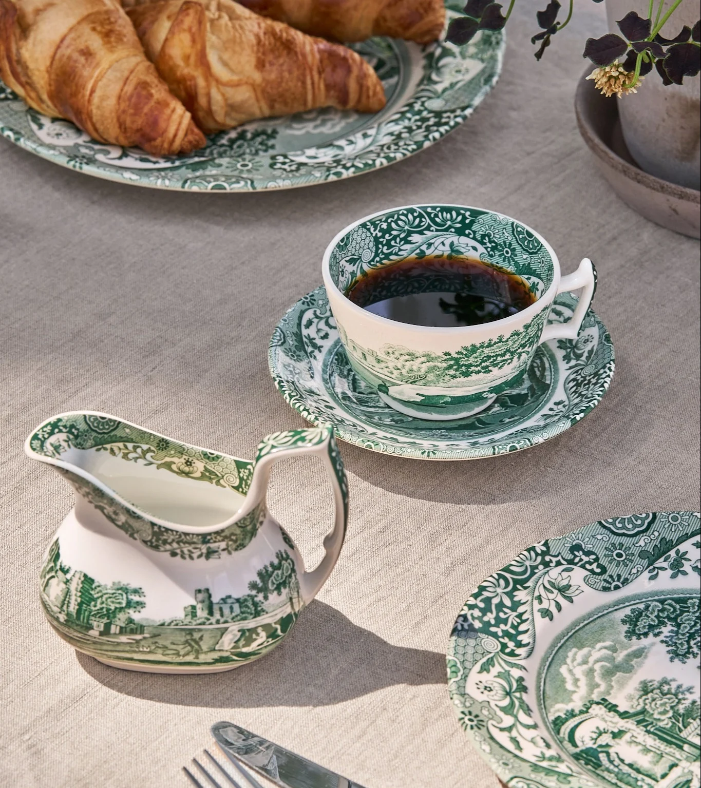 Aamiaispöytä vihreä-valkoisilla kuvioidulla posliinikupilla kahvia, kermakannulla ja lautasilla croissanteja.