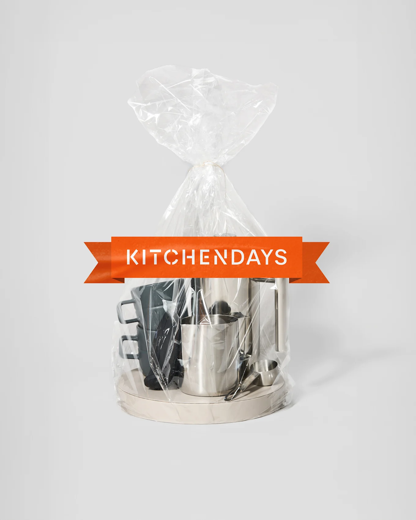 Läpinäkyvä lahjapussi, joka on sidottu narulla, sisältää hopeisen kahvinkeittimen, hopeisen maitokannun ja mustia mukeja vaalealla tarjottimella, oranssilla "KITCHENDAYS"-bannerilla.