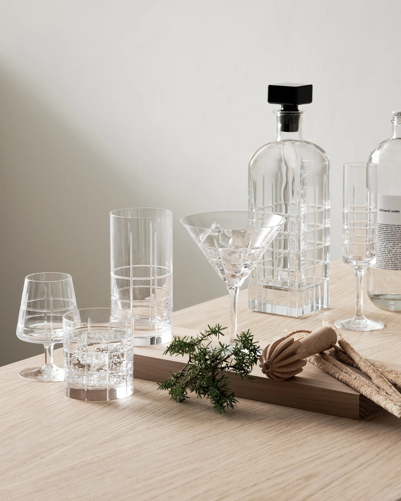 Kirkas lasikaraffi, erilaisia kuvioituja laseja ja kivennäisvesipullo katajanmarjojen ja grissinien kera puupöydällä.