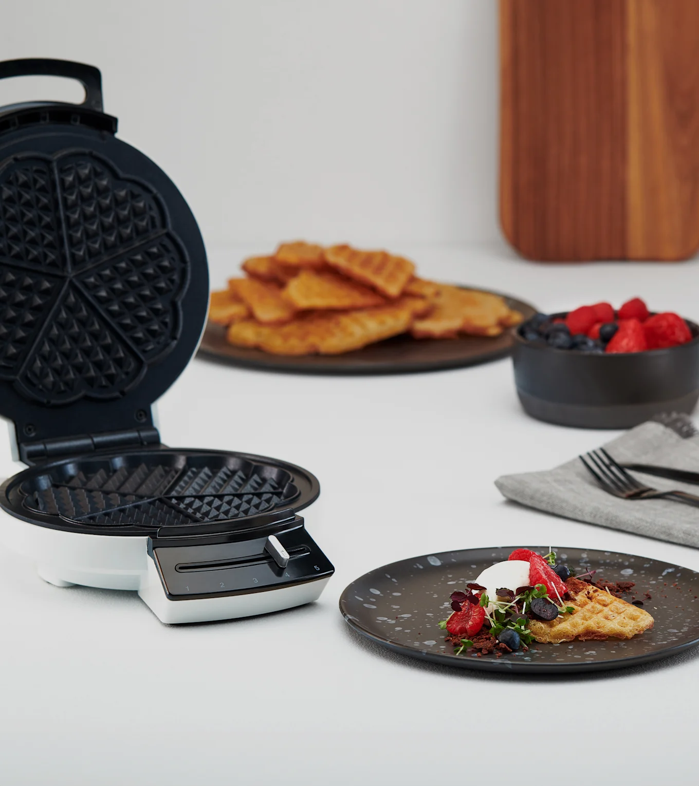 Avoin vohvelirauta lautasen vieressä, jossa vohveli, kermavaahtoa, marjoja ja versoja. Lisää vohveleita ja marjoja taustalla.