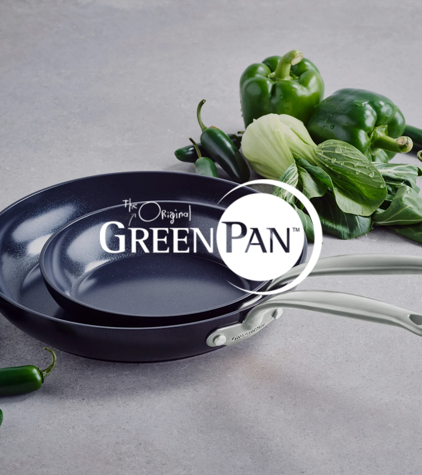 Kaksi tummaa tarttumatonta pannua vaaleilla kahvoilla, pinottuna, vihreiden paprikoiden, paksoin ja jalapeñojen vieressä. GreenPan logo.