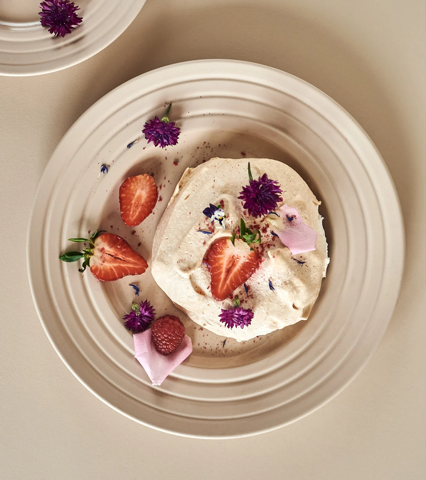 Kermainen jälkiruoka mansikoilla, vadelmalla ja violeteilla kukilla beigellä uurretulla lautasella.