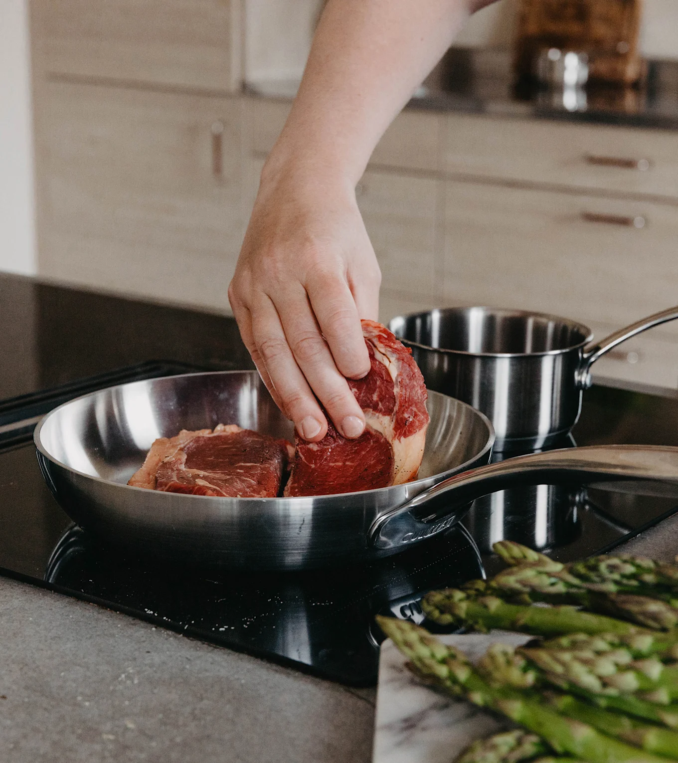Käsi asettaa raa'an pihvin teräspannuun liedellä, parsaa marmorilaudalla etualalla.