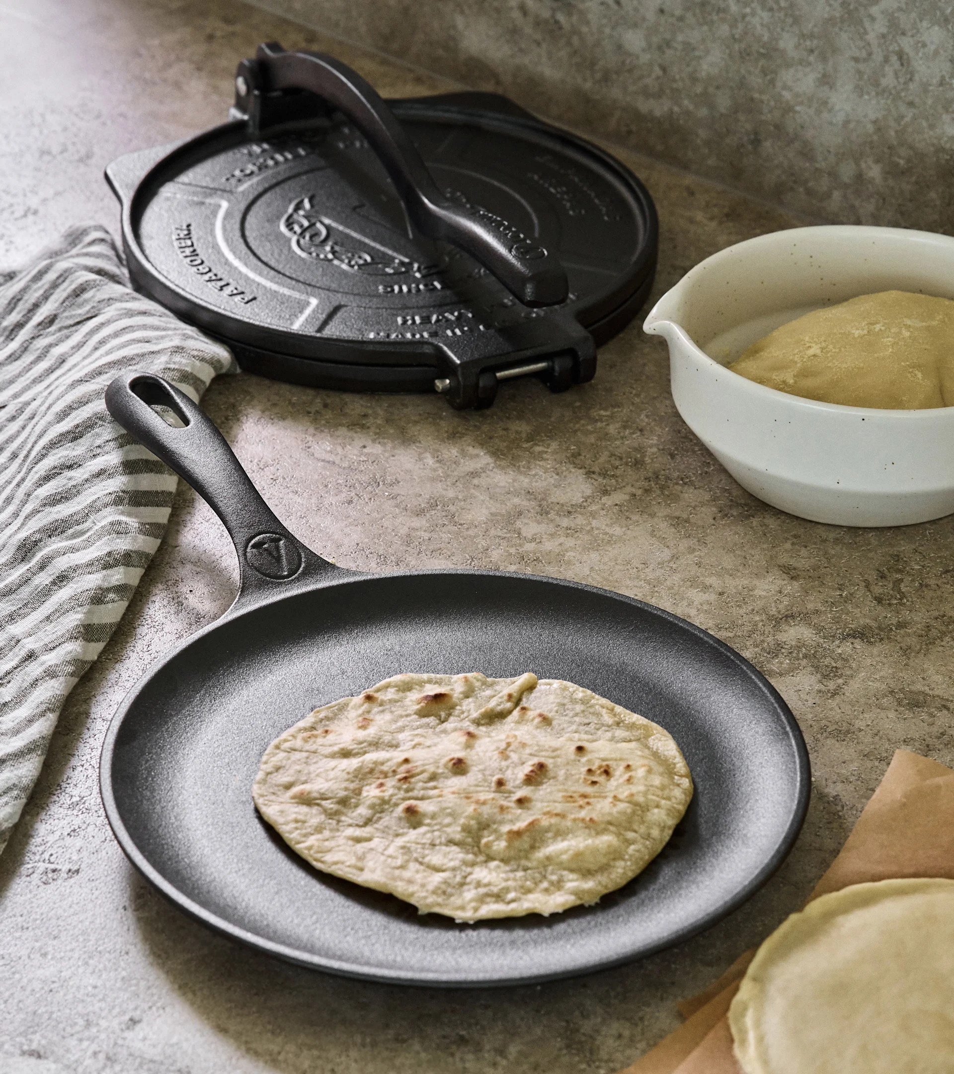 Valurautainen tortillapainin ja pannu kypsennetyllä tortillalla, taikinalla ja raaoilla tortilloilla.