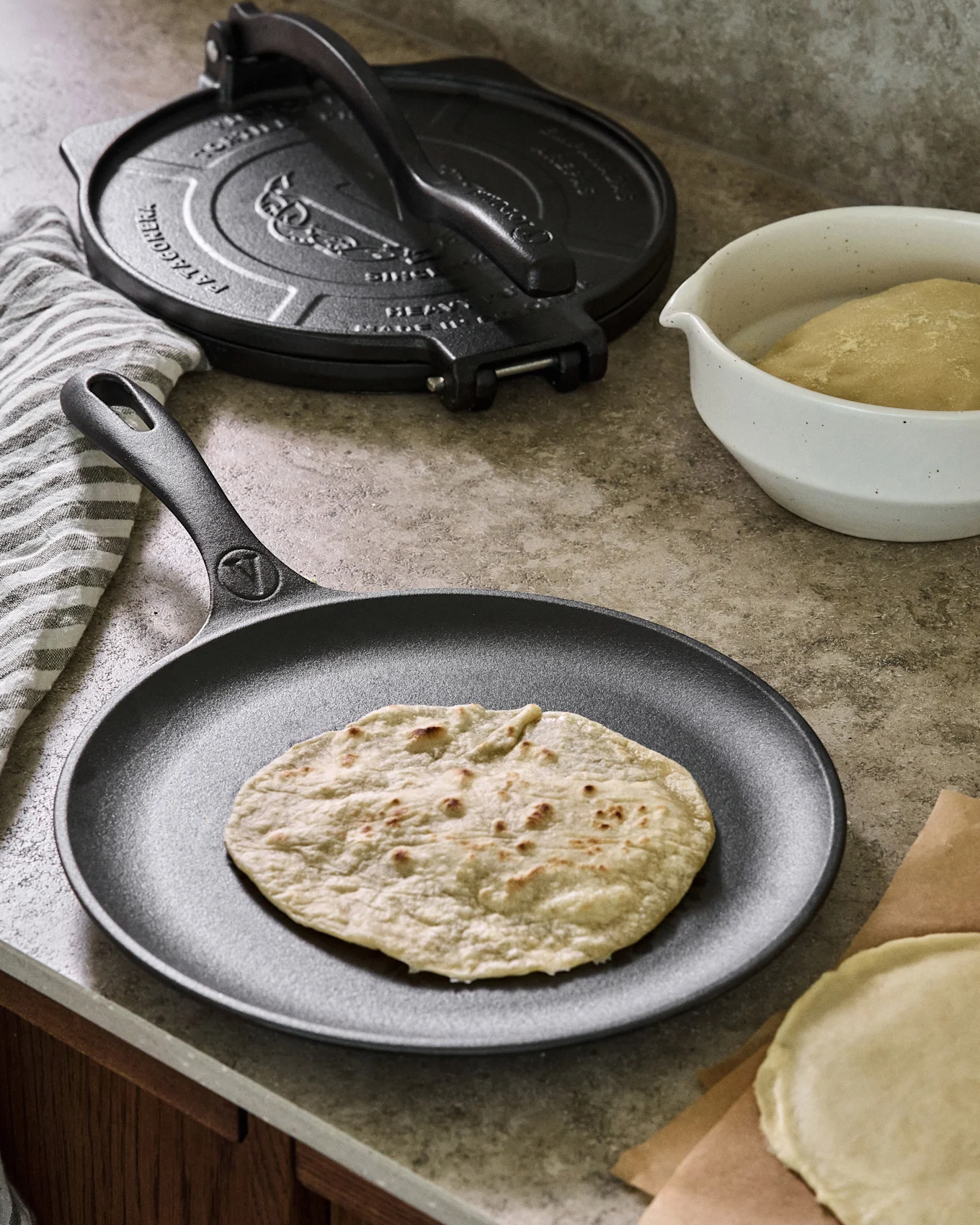 Paistettu tortilla lepää mustalla valurautaparilalla, taustalla tortillapuristin, taikinakulho ja raaka tortilla.