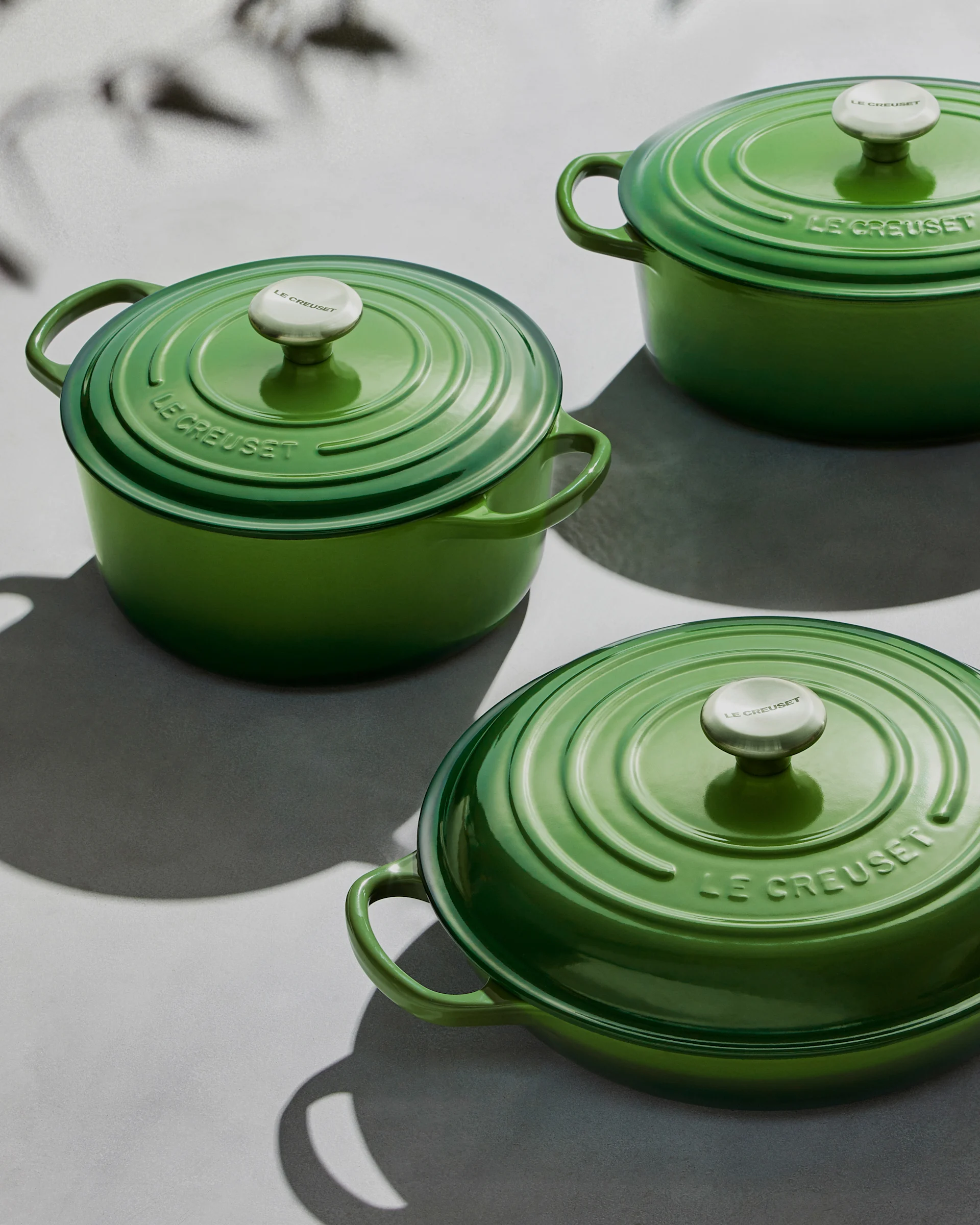 Kolme vihreää Le Creuset -pataa kansineen ja hopeanuppeineen vaalealla pinnalla varjoineen.