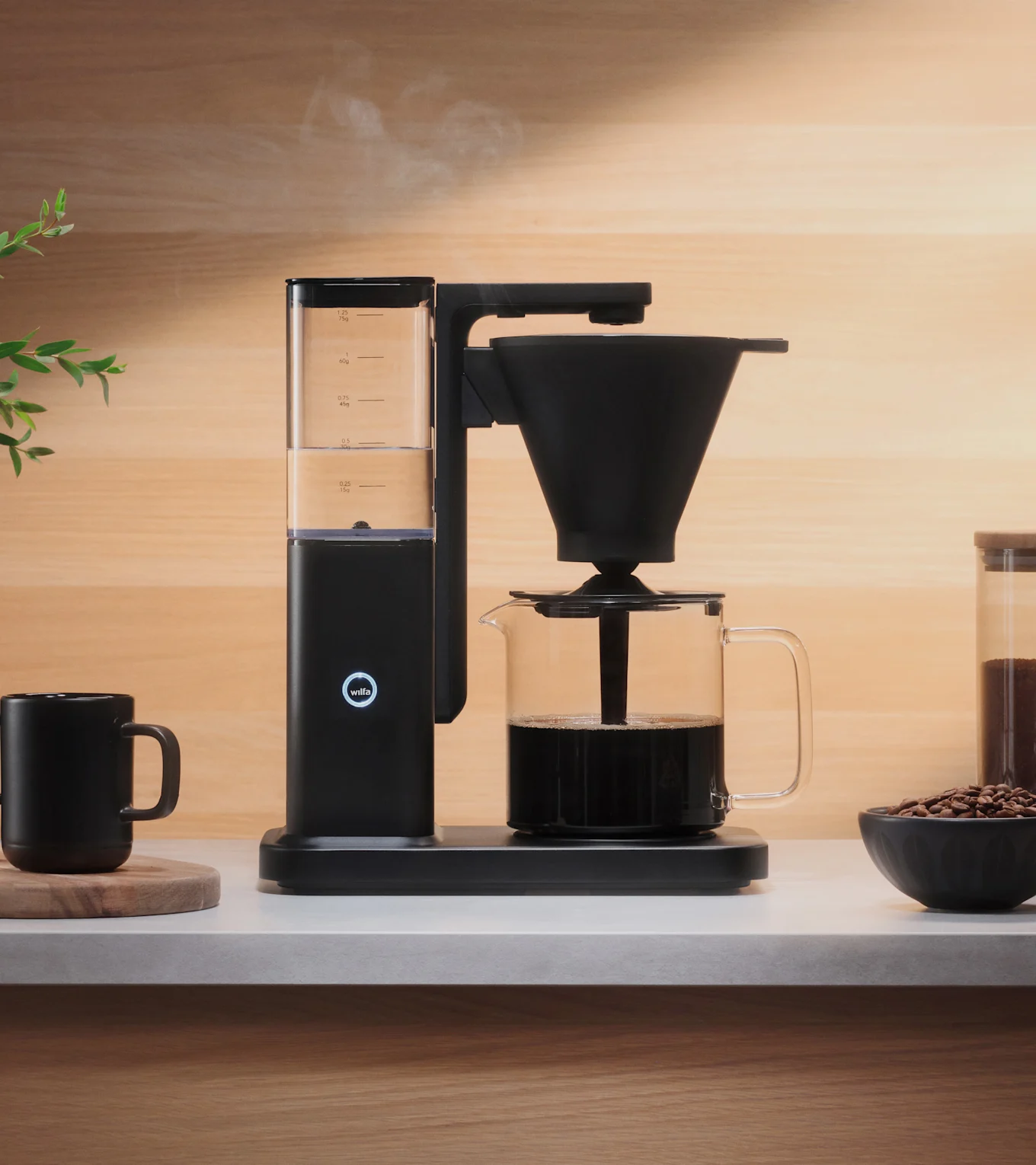 Musta kahvinkeitin valmistaa kahvia lasikannuun vaalealla tiskillä, höyry nousee. Musta muki ja kahvipapuja ovat lähellä.