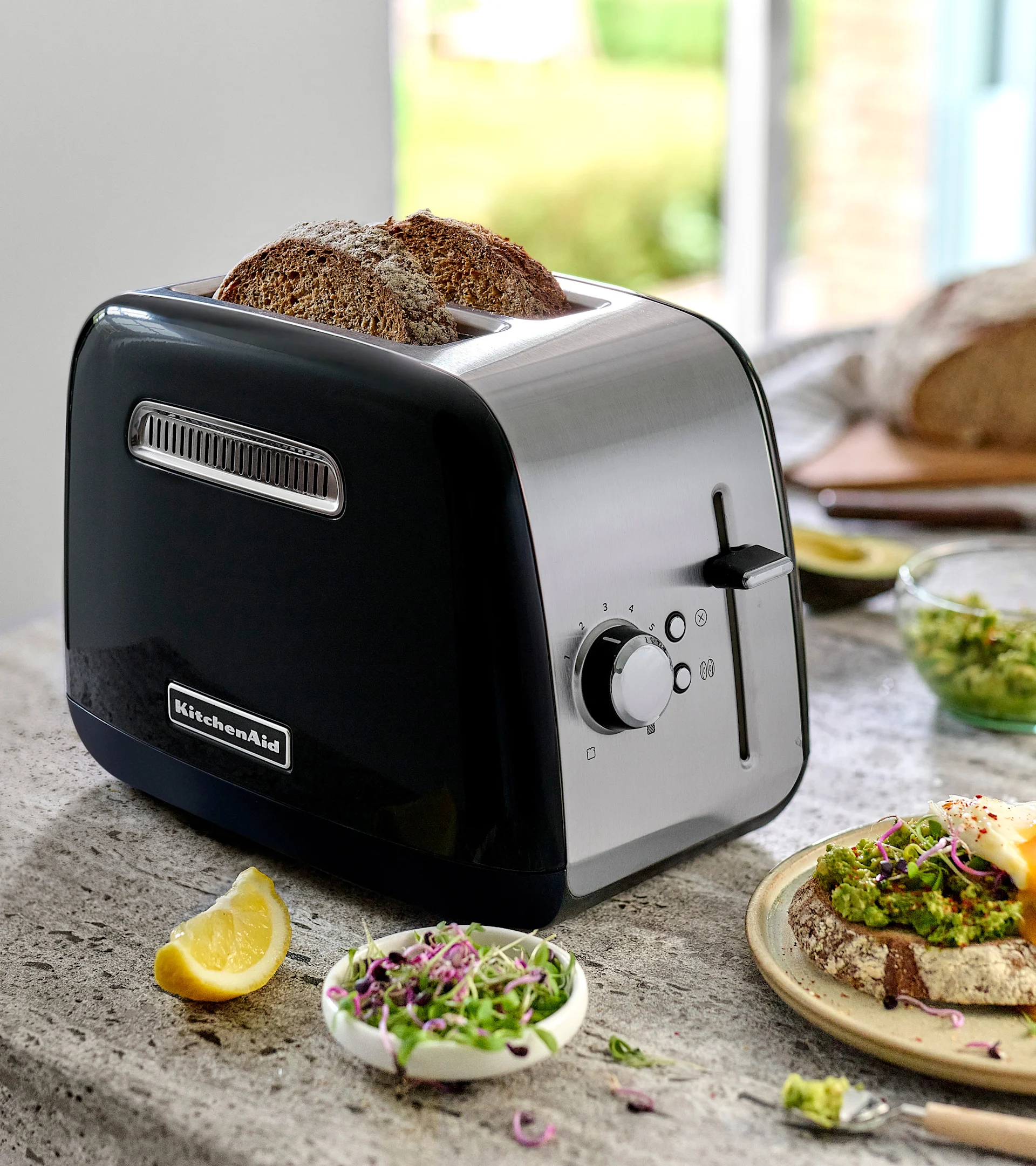 Musta ja ruostumattomasta teräksestä valmistettu KitchenAid leivänpaahdin kahdella tummalla leipäviipaleella. Tiskillä avokadoleipä, kananmuna, ituja ja sitruunaviipale.