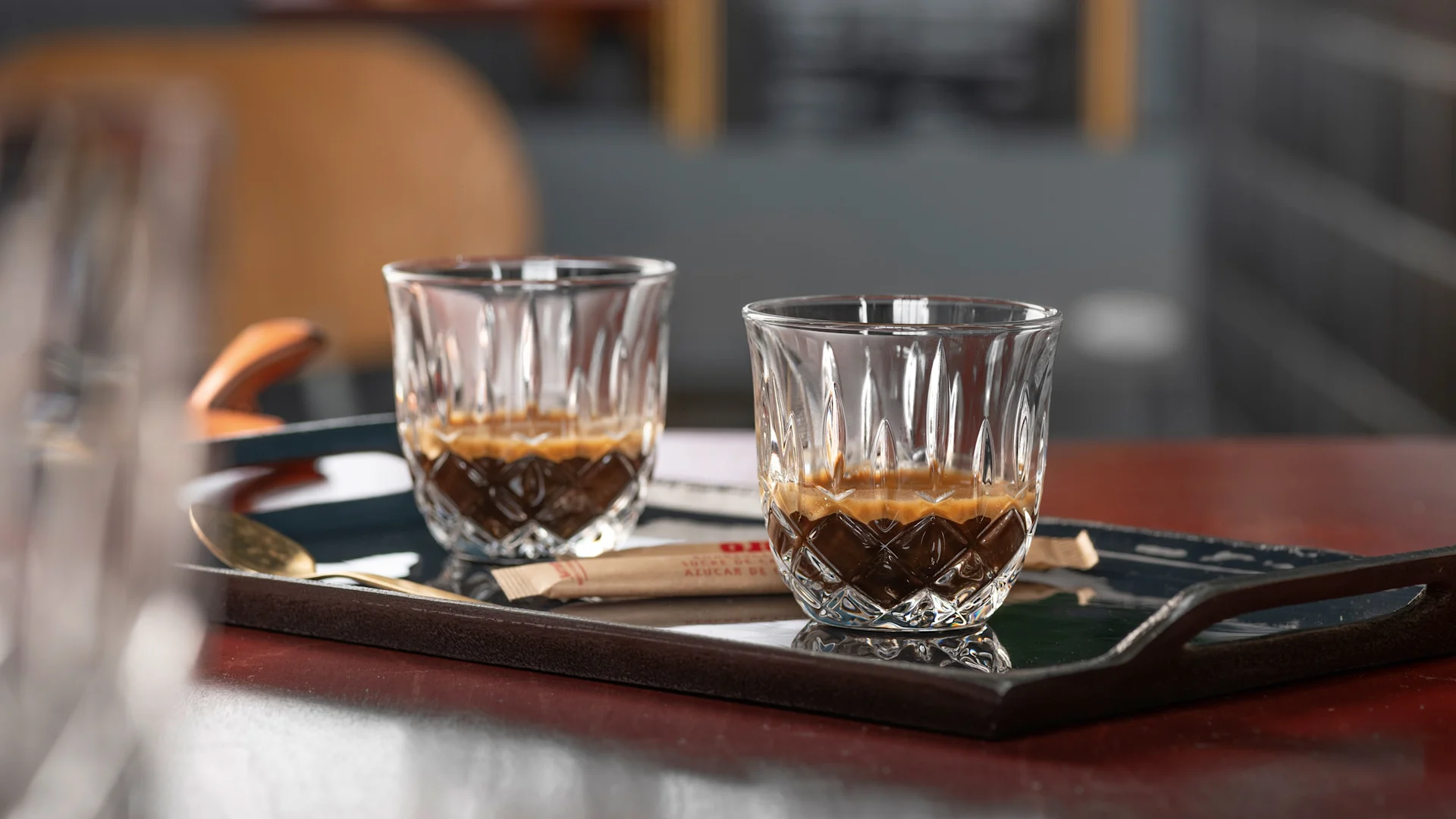 Kaksi koristeellista espressolasia creman kera tummalla tarjottimella, lusikan ja sokeripakkauksen kanssa.