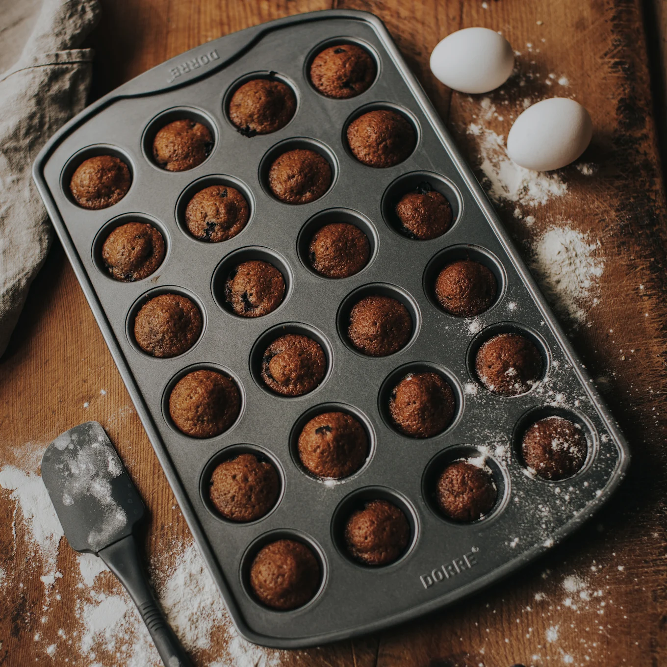 Muffinssivuoka, jossa on 24 paistettua minimuffinssia puupinnalla jauhojen, kahden valkoisen munan ja lastan kera.