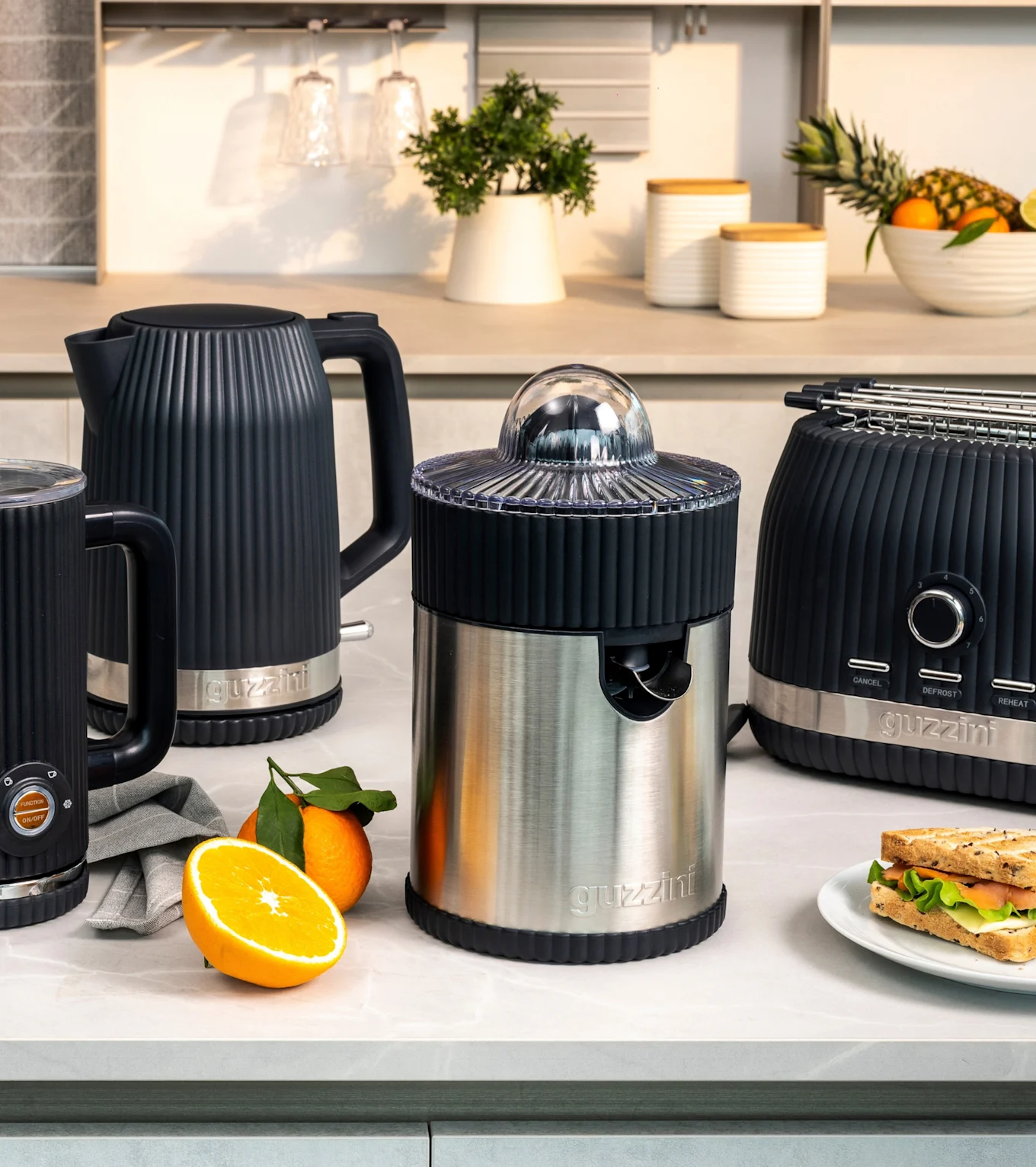 Musta uurrettu vedenkeitin, maidonvaahdotin, teräsmehustin ja leivänpaahdin appelsiinien ja voileivän kera.