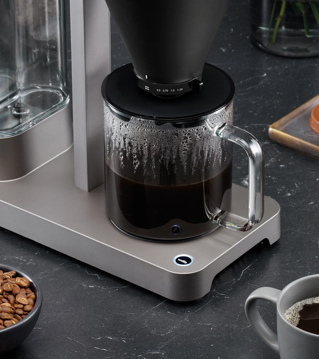 Moderni kahvinkeitin mustalla suodatinosalla valmistaa kahvia lasikannuun, tummalla työtasolla, kahvipapuja ja muki.