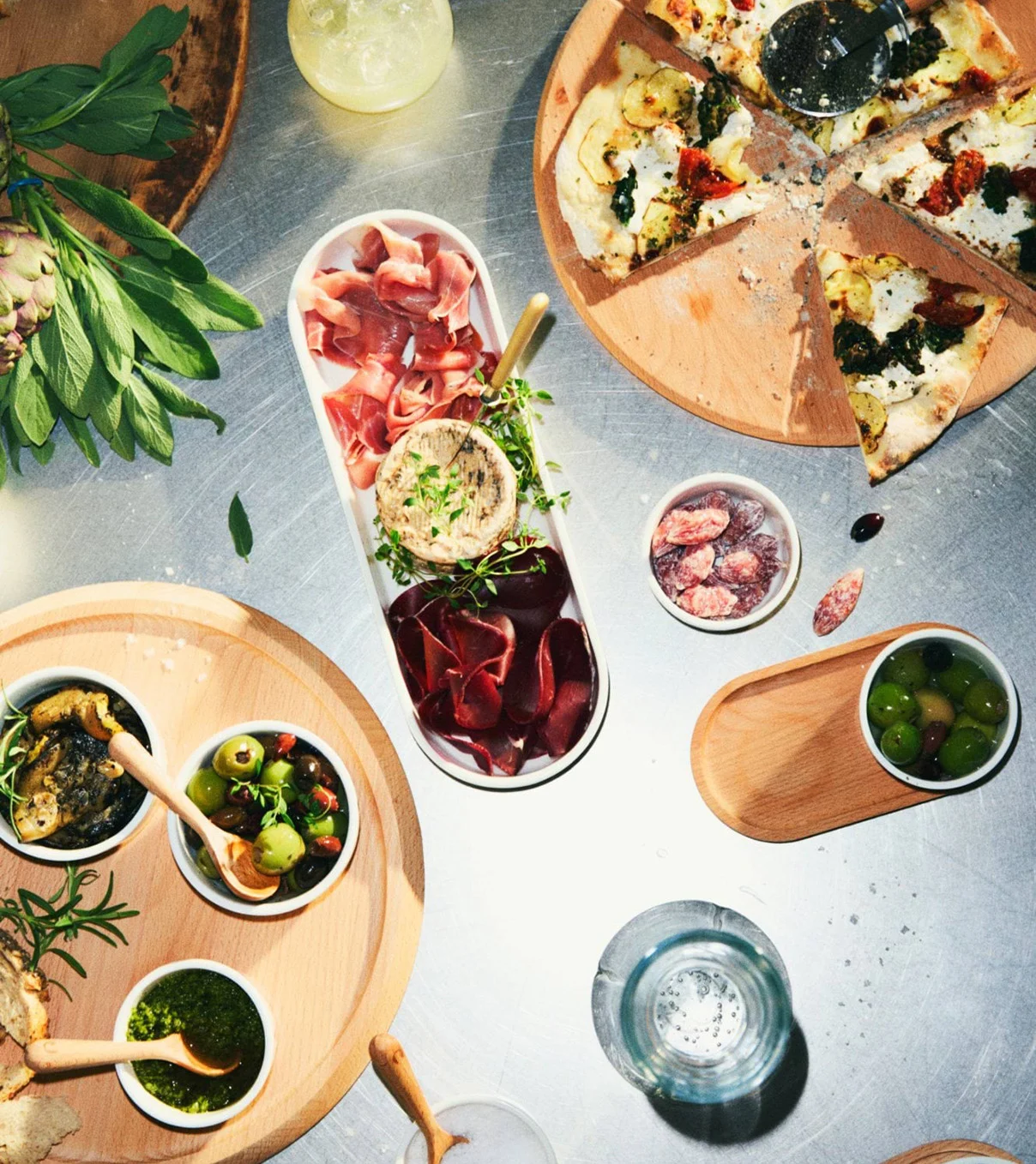 Yleiskuva ruokapöydästä, jossa on viipaloitu pizza, leikkeleitä, oliiveja, pestoa ja tapenadea puulaudoilla.