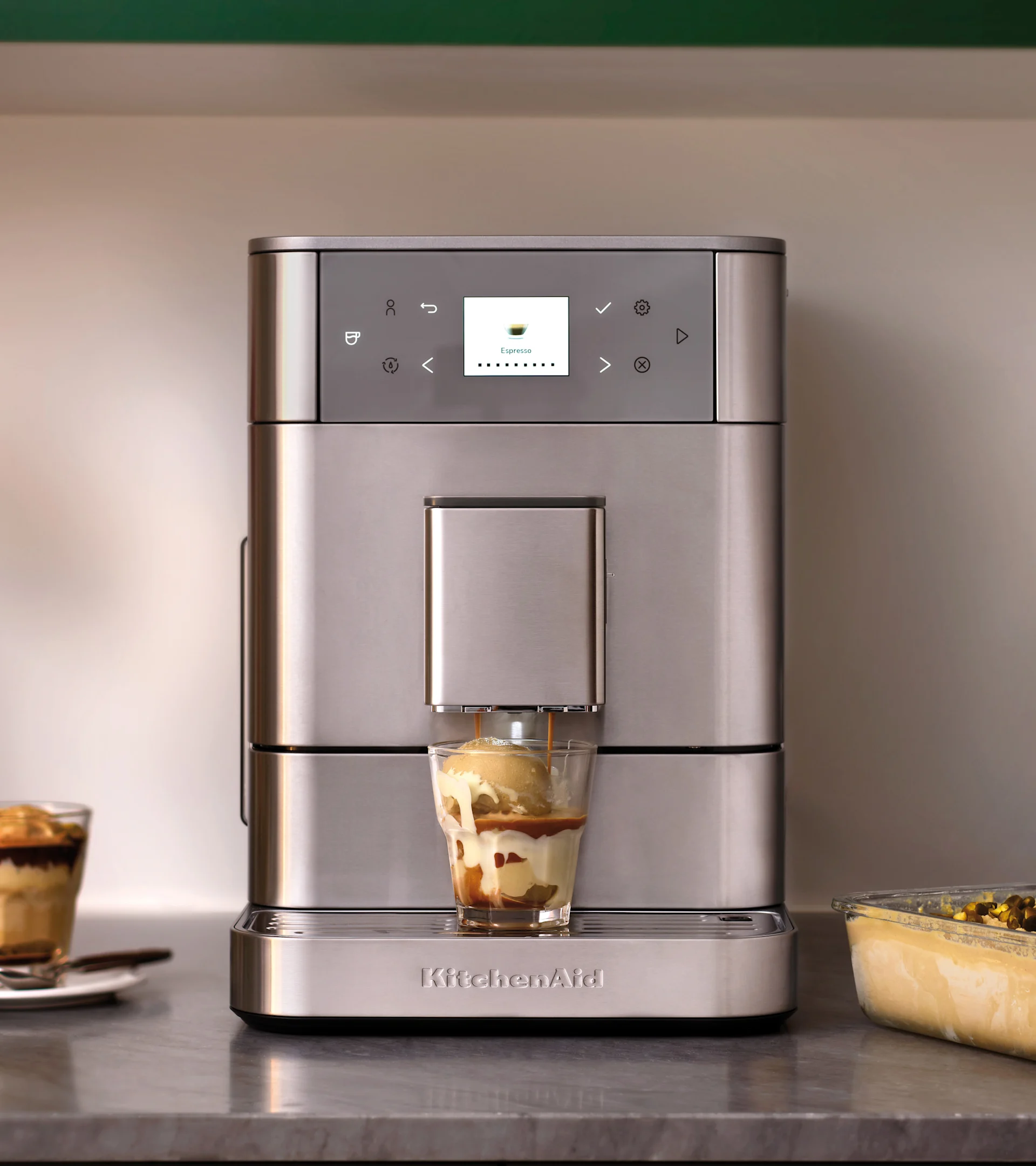 Hopeinen KitchenAid espressokone annostelee espressoa jäätelölasille, valmistaen affogaton. Toinen jälkiruoka taustalla.