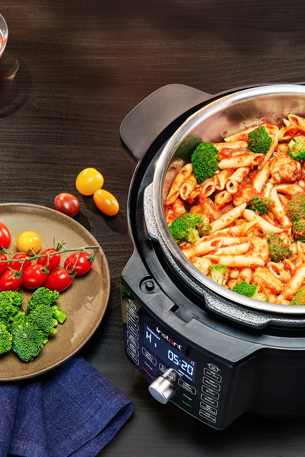 Musta monitoimikeitin, jossa on penne-pastaa ja parsakaalia tomaattikastikkeessa, vieressään lautanen tuoreita kirsikkatomaatteja ja parsakaalia tummalla puupöydällä.